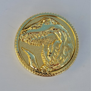 May include: Gold-colored metal coin with a raised image of a Tyrannosaurus Rex dinosaur head. The coin has a textured border.