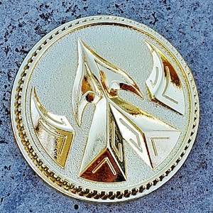 May include: A gold-colored metal coin with a raised design of an arrow with a stylized, geometric pattern. The coin has a textured, circular border.