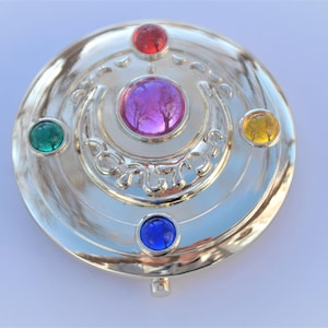 May include: Silver-colored compact mirror with a crescent moon design and five colorful gems: green, red, purple, blue, and yellow.