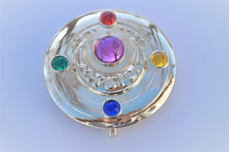 May include: Silver-colored compact mirror with a crescent moon design and five colorful gems: green, red, purple, blue, and yellow.
