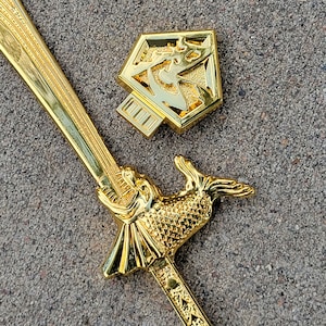 May include: A gold-colored sword with a fish-shaped handle and a decorative design on the blade. The sword is lying on a gray surface next to a gold-colored hexagonal pendant with a geometric design.