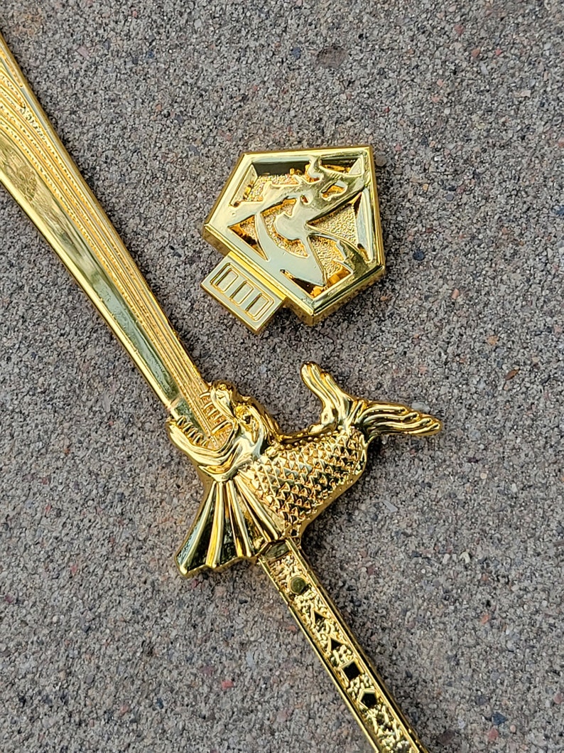 May include: A gold-colored sword with a fish-shaped handle and a decorative design on the blade. The sword is lying on a gray surface next to a gold-colored hexagonal pendant with a geometric design.
