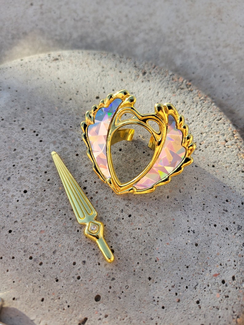 May include: A gold-tone heart-shaped ring with iridescent pink and blue accents. The ring is accompanied by a gold-tone sword-shaped charm with a clear rhinestone.