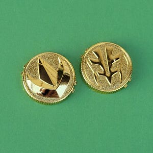 May include: Two gold-colored, circular coins with detailed designs. One coin features a stylized bird or wing shape, while the other has a different emblem. The coins are set against a solid green background.