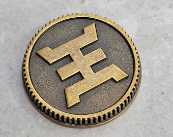Könnte beinhalten: Eine goldfarbene Metallmünze mit einem stilisierten Buchstaben 'H' auf einem dunkelbraunen Hintergrund. Die Münze hat einen zahnradartigen Rand.