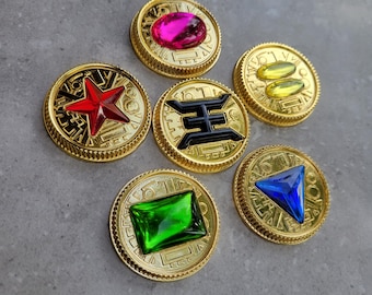 Könnte beinhalten: Sechs goldfarbene Token mit verschiedenen farbigen Edelsteinen in der Mitte. Die Edelsteine sind rot, pink, gelb, grün, blau und schwarz. Jedes Token hat ein einzigartiges Design am Rand.