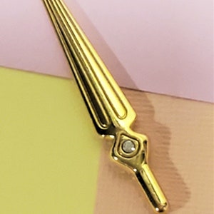 May include: A gold-colored sword pin with a clear rhinestone at the base. The pin is on a pink and yellow background.
