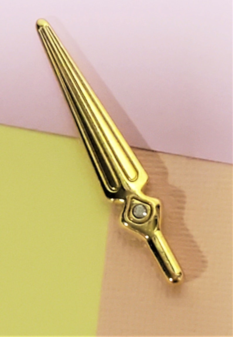May include: A gold-colored sword pin with a clear rhinestone at the base. The pin is on a pink and yellow background.