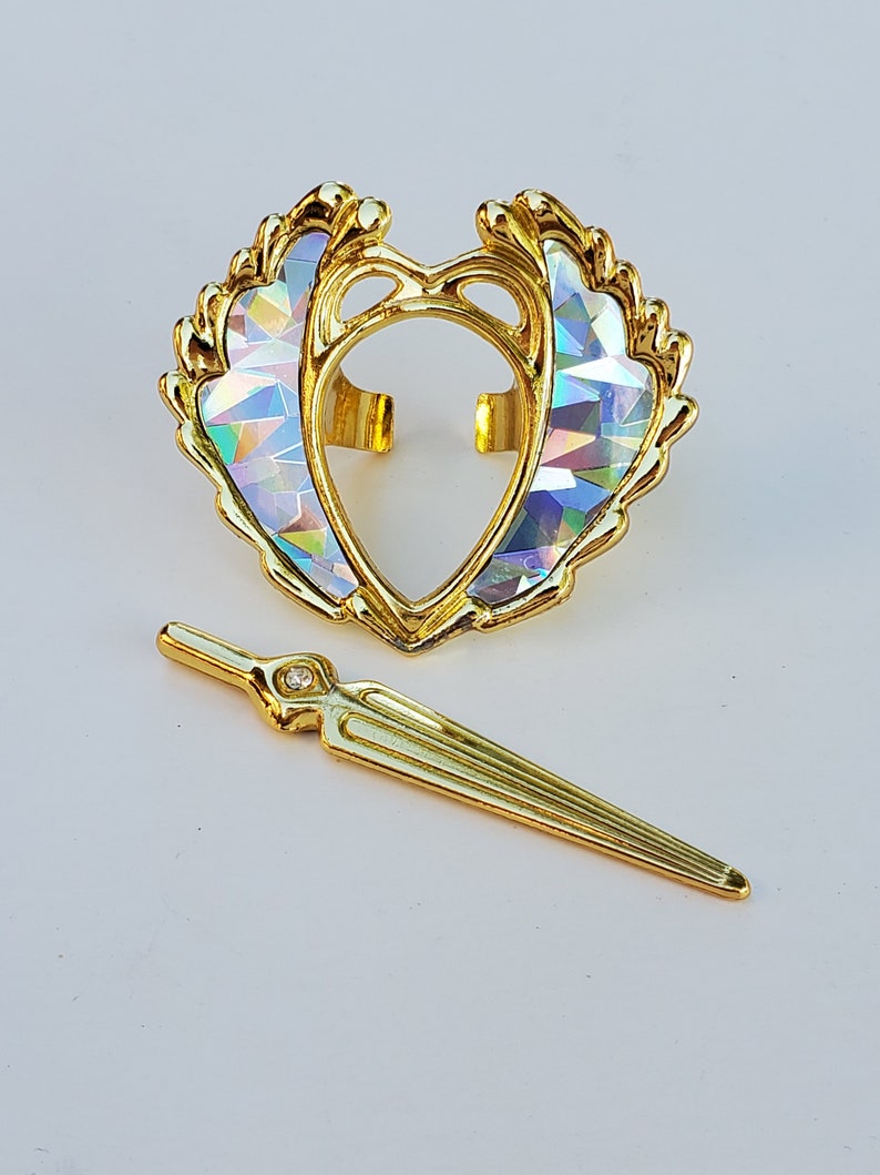 May include: A gold-colored heart-shaped crown with iridescent detailing and a matching gold-colored sword with a single clear rhinestone.