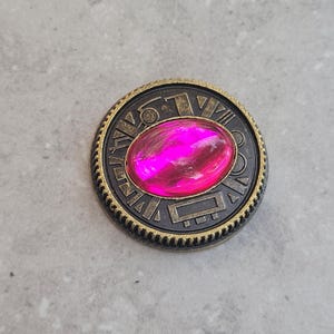May include: A round, gold-colored metal pin with a pink gem in the center. The pin has an intricate design around the gem, including Roman numerals.