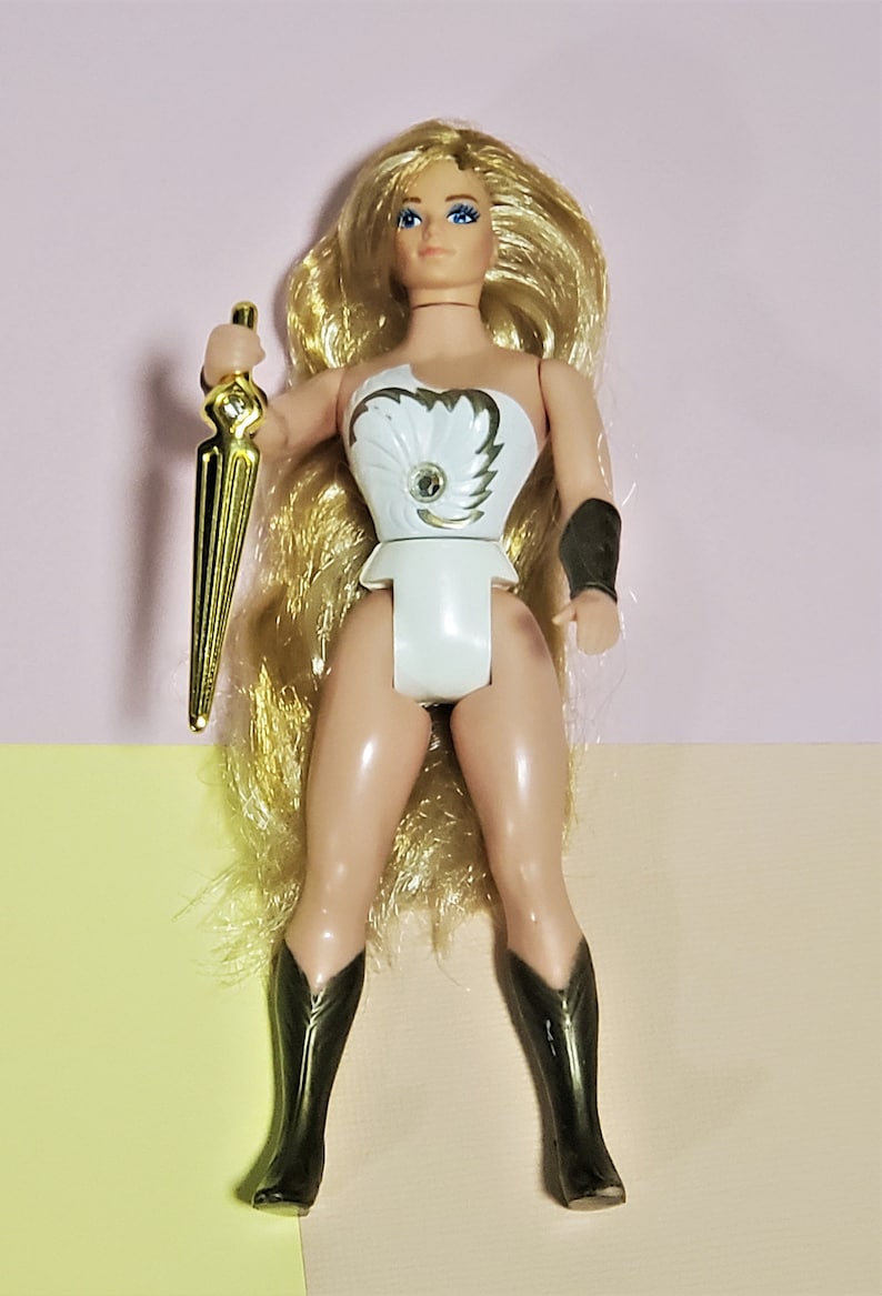 May include: A vintage action figure of a female character with long blonde hair, wearing a white and gold costume and holding a gold sword. She is wearing black boots.