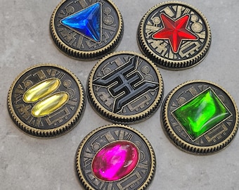 Könnte beinhalten: Sechs runde, goldfarbene Metallmarken mit verschiedenen farbigen Edelsteinen und Symbolen in der Mitte. Die Edelsteine sind blau, rot, gelb, grün und pink. Die Symbole sind ein Dreieck, ein Stern, zwei Ovale, ein Buchstabe H und ein Quadrat.