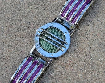 May include: A silver bracelet with a purple enamel band and a round, silver, decorative element with a green mesh screen.