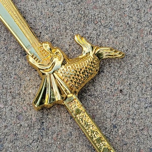 May include: A gold-colored sword with a fish-shaped handle. The handle is detailed with scales and the sword has a decorative pattern.