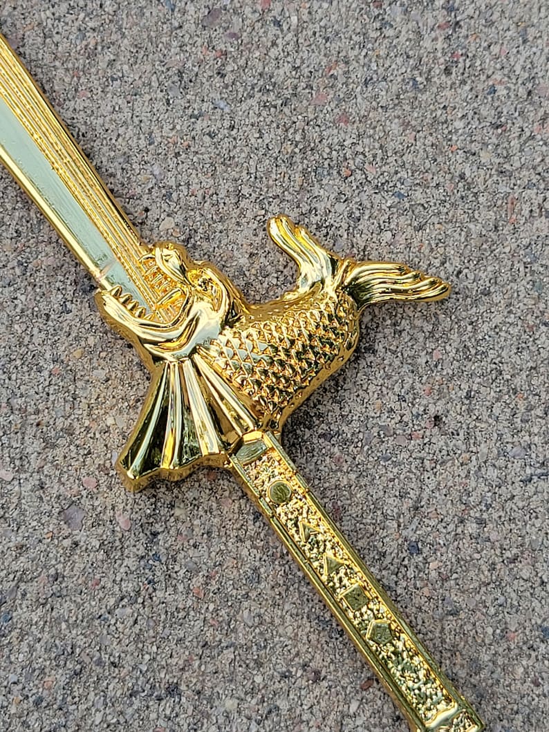 May include: A gold-colored sword with a fish-shaped handle. The handle is detailed with scales and the sword has a decorative pattern.