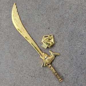 May include: A gold-colored decorative sword with a curved blade and a fish-shaped handle. The sword is on a gray textured surface.