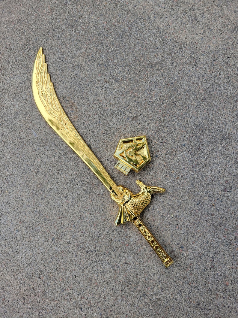 May include: A gold-colored decorative sword with a curved blade and a fish-shaped handle. The sword is on a gray textured surface.