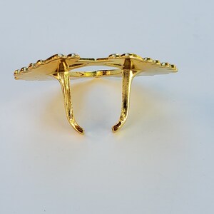 May include: Gold-colored metal ring holder with a decorative design. The holder has two prongs that extend upwards.