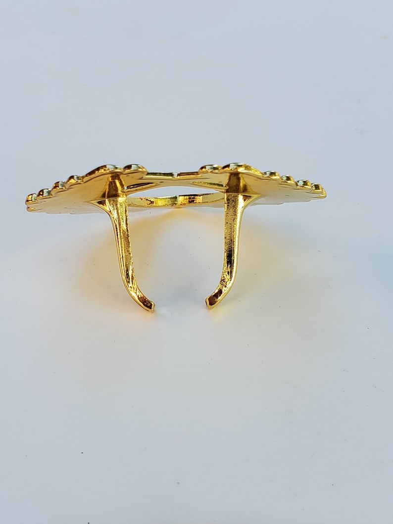May include: Gold-colored metal ring holder with a decorative design. The holder has two prongs that extend upwards.