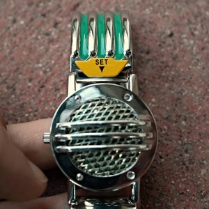 K&ouml;nnte beinhalten: Eine silberne und gr&uuml;ne Armbanduhr mit einem runden Zifferblatt und einem Gittermuster. Die Uhr hat einen gelben Knopf mit dem Wort "SET" darauf.