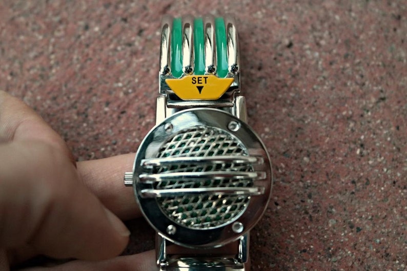 K&ouml;nnte beinhalten: Eine silberne und gr&uuml;ne Armbanduhr mit einem runden Zifferblatt und einem Gittermuster. Die Uhr hat einen gelben Knopf mit dem Wort "SET" darauf.