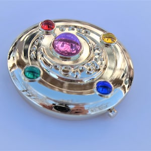 May include: A gold-colored compact mirror with a crescent moon design and five colorful gemstones. The gemstones are red, green, blue, yellow, and purple.