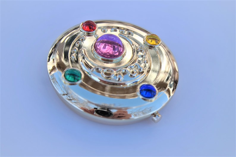 May include: A gold-colored compact mirror with a crescent moon design and five colorful gemstones. The gemstones are red, green, blue, yellow, and purple.