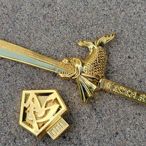May include: A gold-colored sword with a dragon head handle and a decorative sheath. The sword is lying on a gray concrete surface.