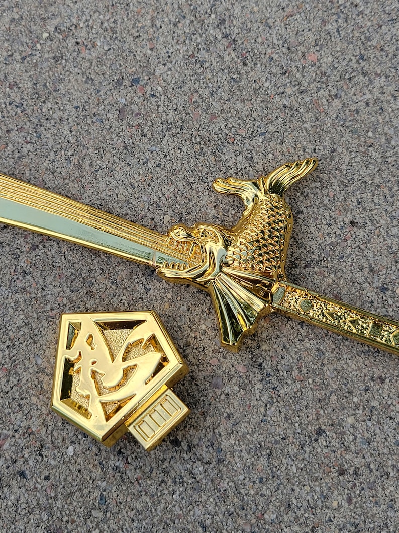 May include: A gold-colored sword with a dragon head handle and a decorative sheath. The sword is lying on a gray concrete surface.