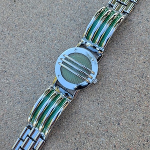 May include: A silver-tone wristwatch featuring a circular case with a green mesh dial protected by three horizontal silver bars. The band consists of alternating silver-tone metal links and curved, glossy green segments that match the dial's color.