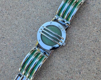 May include: A silver-tone wristwatch featuring a circular case with a green mesh dial protected by three horizontal silver bars. The band consists of alternating silver-tone metal links and curved, glossy green segments that match the dial's color.