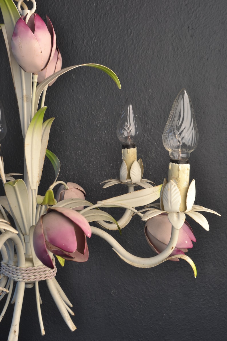 Pu&ograve; includere: Lampadario in metallo bianco con accenti di fiori rosa e bianchi e lampadine in vetro trasparente. Il lampadario ha un design floreale con foglie e fiori.