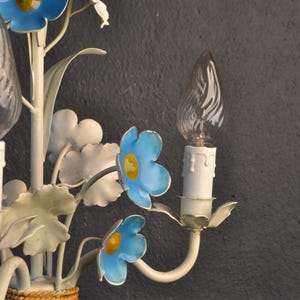 May include: A white metal wall sconce with blue and yellow flowers and clear glass bulbs. The sconce has a woven brown band around the base.