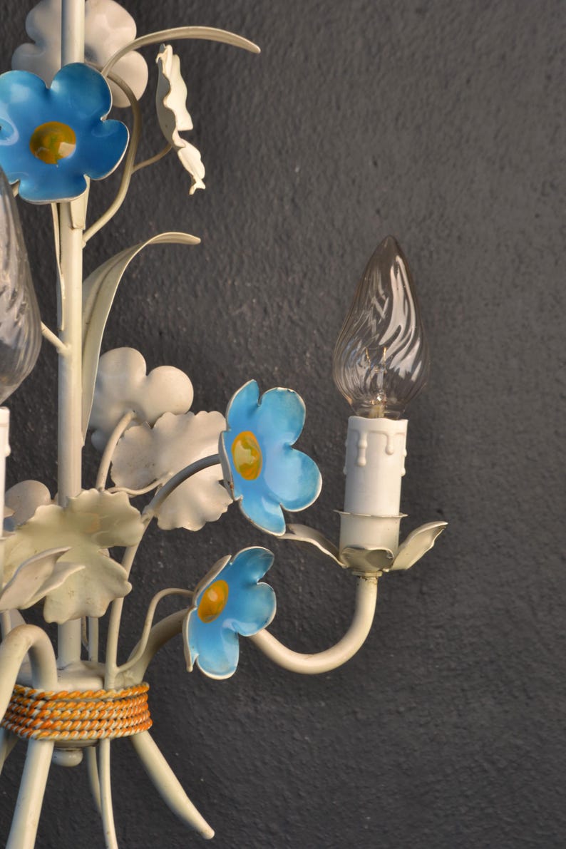 May include: A white metal wall sconce with blue and yellow flowers and clear glass bulbs. The sconce has a woven brown band around the base.