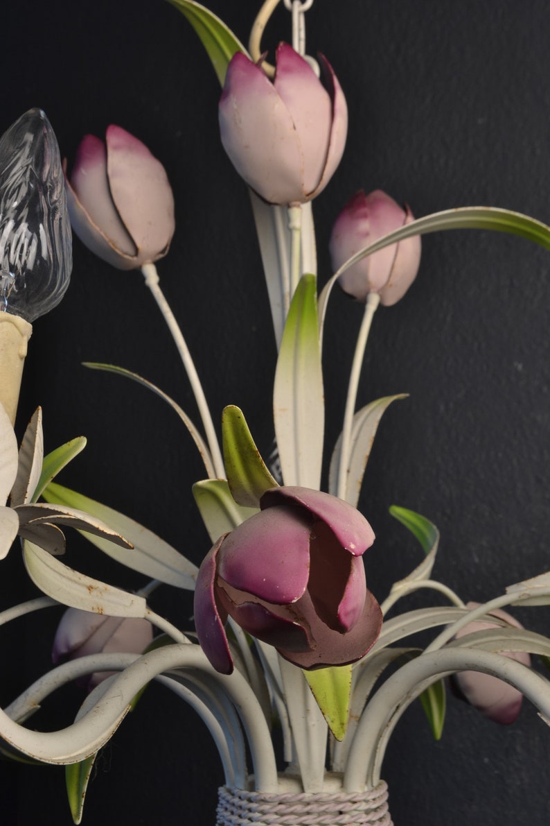 Pu&ograve; includere: Un lampadario in metallo bianco con fiori a forma di tulipano rosa e viola. Il lampadario ha una base in corda bianca.