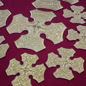 May include: A collection of gold-colored, intricately designed, cross-shaped ornaments displayed on a red velvet surface.