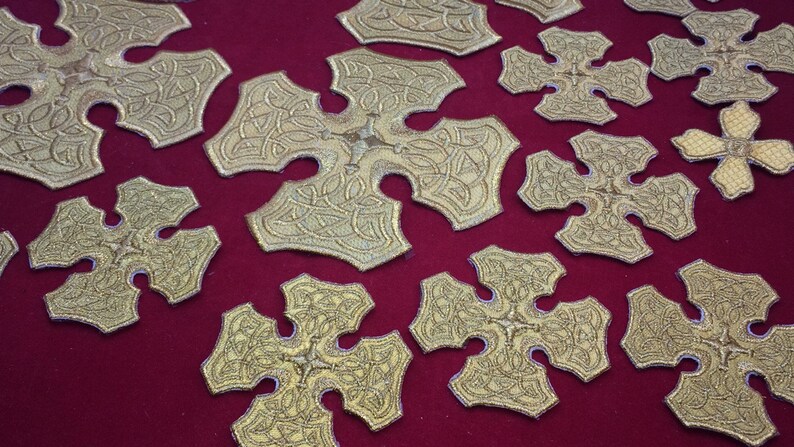 May include: A collection of gold-colored, intricately designed, cross-shaped ornaments displayed on a red velvet surface.
