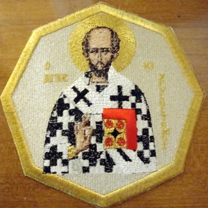 May include: Embroidered patch with a gold border, depicting a religious icon of a man with a beard, wearing a white and black chequered robe, holding a red book. The icon is surrounded by a gold halo and the text "O Brave" and "O Excellent" is visible on the patch.