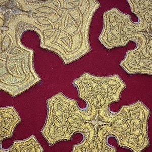 May include: A close-up of a red fabric background with multiple gold embroidered crosses. The crosses have a Celtic knot design.