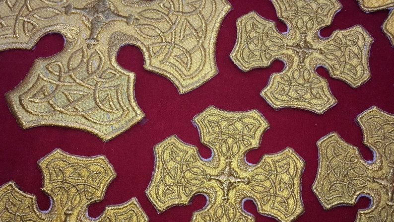 May include: A close-up of a red fabric background with multiple gold embroidered crosses. The crosses have a Celtic knot design.