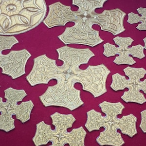 May include: A collection of gold embroidered crosses with intricate designs. The crosses are arranged on a red fabric background.
