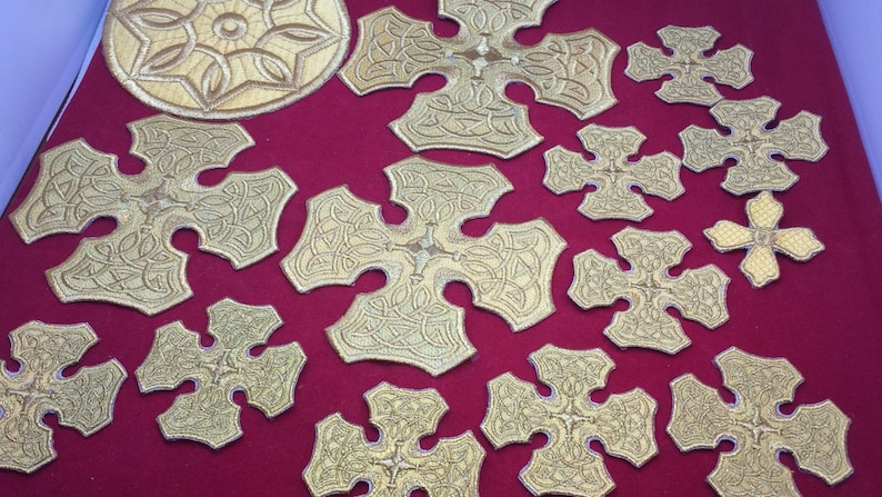 May include: A collection of gold embroidered crosses with intricate designs. The crosses are arranged on a red fabric background.