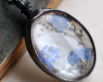 May include: An oval pendant necklace featuring a dark metal frame and chain. The pendant contains a clear, rounded glass insert displaying pressed blue forget-me-not flowers and delicate white wisps against a light, neutral background.