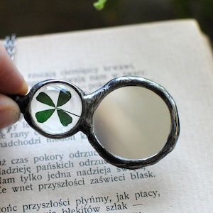 May include: A silver necklace with a magnifying glass charm and a four-leaf clover encased in glass. The necklace is lying on a book with text in Polish.
