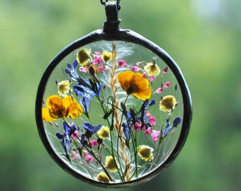 Puede incluir: Un delicado collar con un colgante de vidrio transparente que presenta flores silvestres prensadas en tonos de amarillo, azul y rosa.