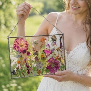 Könnte beinhalten: Ein rechteckiger Glasrahmen mit einem dünnen Metallrand, der an einer feinen Metallkette hängt. Der Rahmen enthält ein gepresstes Arrangement aus getrockneten Wildblumen, darunter große rosa und violette Blüten, kleine orangefarbene Knospen.