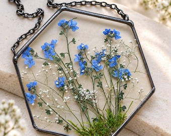 May include: A hexagonal pendant featuring pressed blue forget-me-not flowers, white baby's breath, and green moss encased in clear glass with a dark metal frame. The pendant hangs from a matching dark metal chain link necklace.