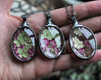 Può includere: Tre collane con ciondoli ovali in argento, con fiori pressati incastonati in resina trasparente. I ciondoli presentano un mix di fiori bianchi, rosa e verdi.