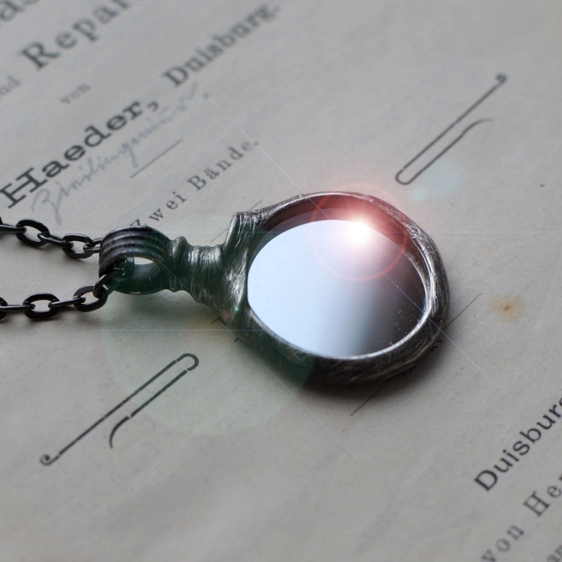 May include: A circular pendant with a dark, textured metallic frame and a reflective center, attached to a dark chain. The pendant features a prominent, bright light flare on its surface, creating a high-contrast visual effect against the dark metal.