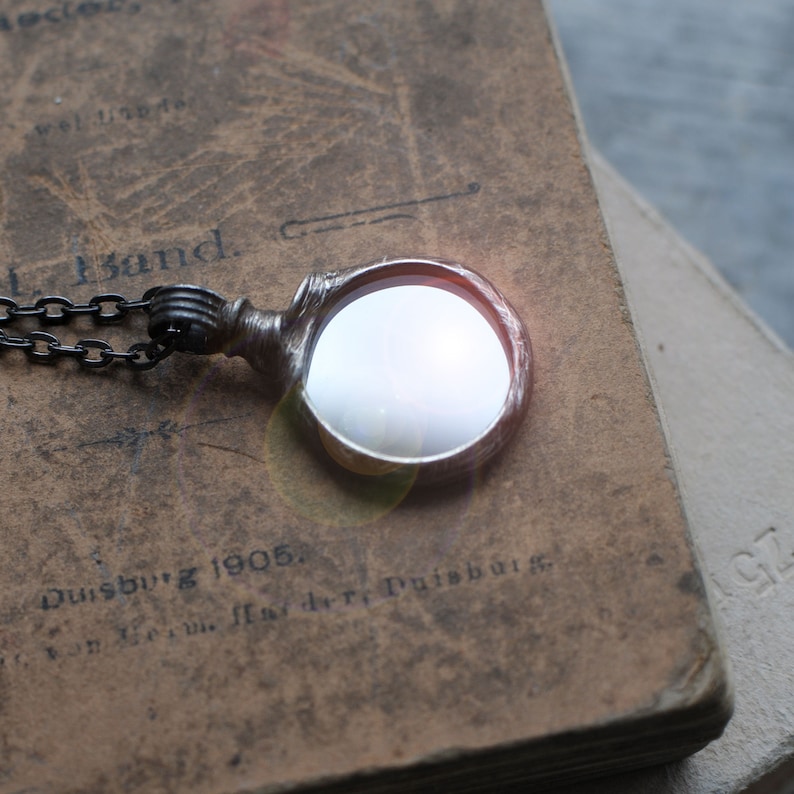 May include: A circular magnifying glass pendant with a metallic frame and a dark chain. The lens is clear and reflective, showing a bright light reflection. The pendant is resting on a weathered, brown, vintage-style book cover with visible text including the word Duisburg 1905.